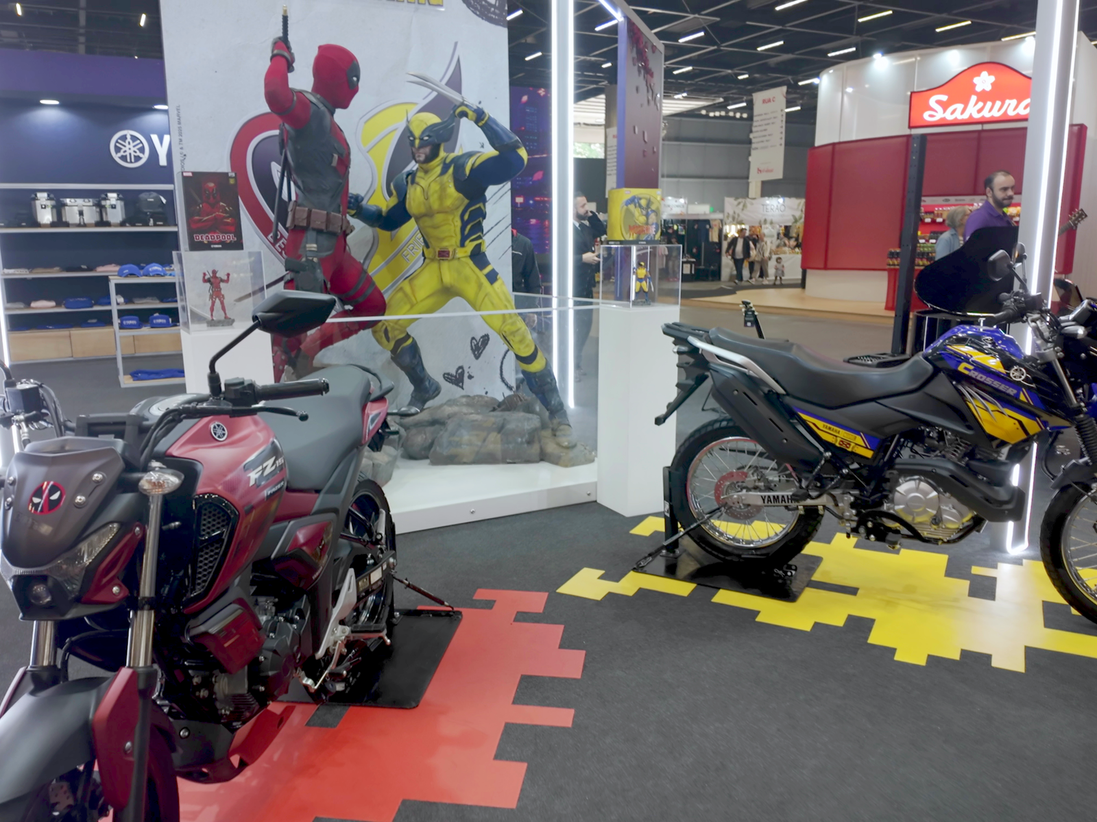 Motos, uma amarela e uma vermelha, ao fundo duas estátuas em tamanho real dos heróis Deadpool e Wolverine.